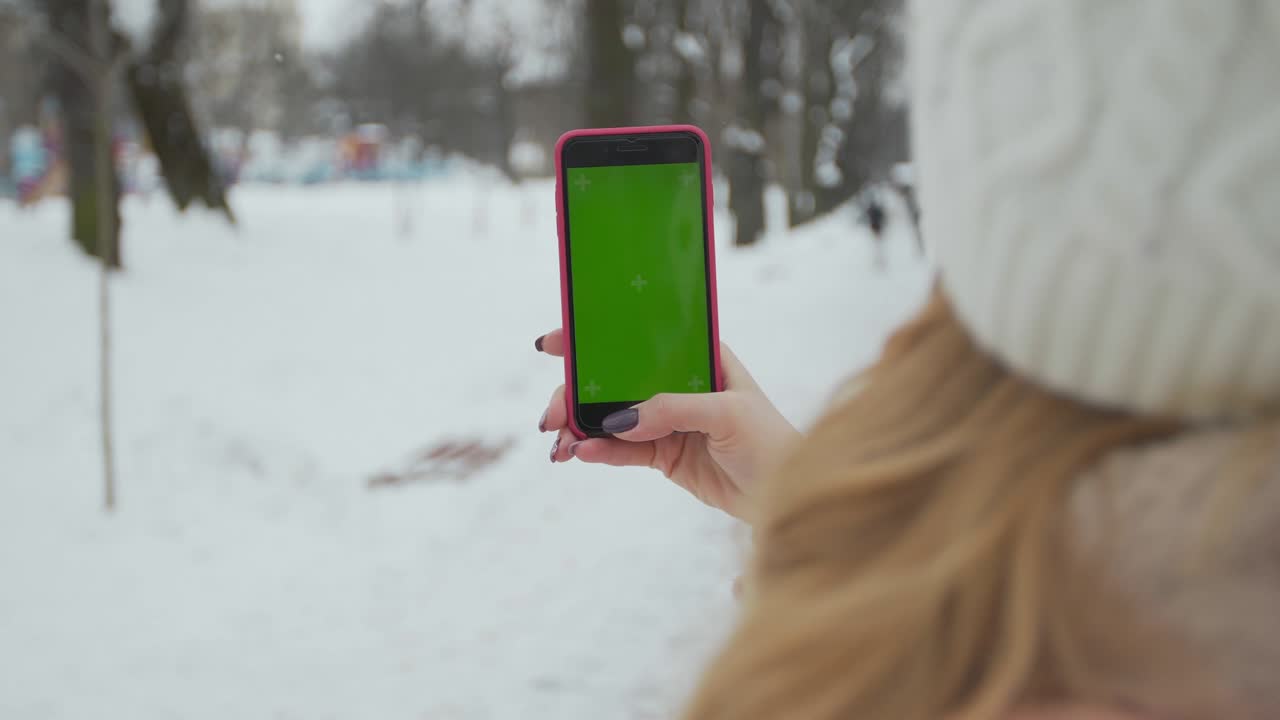 una mujer joven escribe y desliza en su teléfono inteligente en el parque de invierno. teléfono celular con pantalla de teclas cromadas - pantalla verde. cerrar. seguimiento de movimiento.