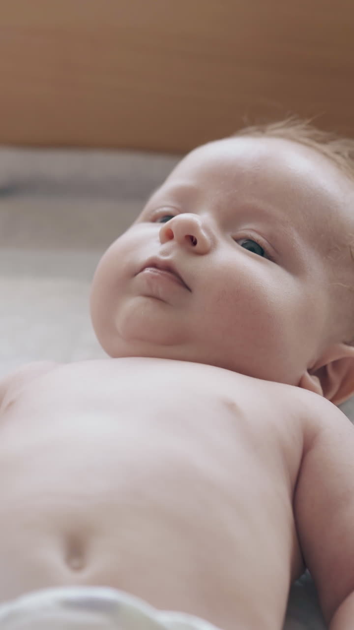 serious newborn boy with short fair hair and in white diaper lies on baby changing table and moves body close view