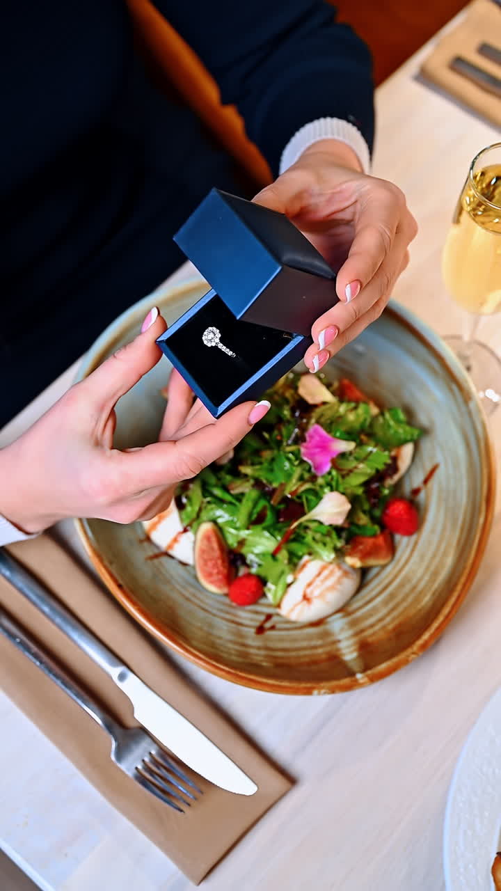 Hænder, der holder et smykkeskrin over en salat og en drink ved et spisebord. En person holder et åbent smykkeskrin over en salat og et glas drink på et bord.