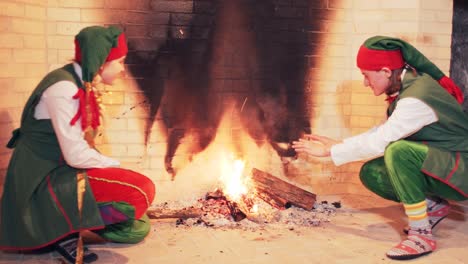 elves in green fairytale costumes are sitting by a large fireplace with fire. christmas atmosphere indoors