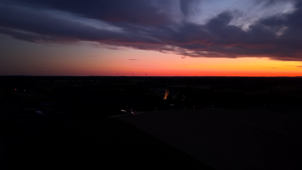 Dramatische wolken aan de oranje hemel in de ochtend. Verlichte kerktoren van een kleine stad en rijdende auto's. Brede luchtfoto. Vredige scène bij zonsopgang