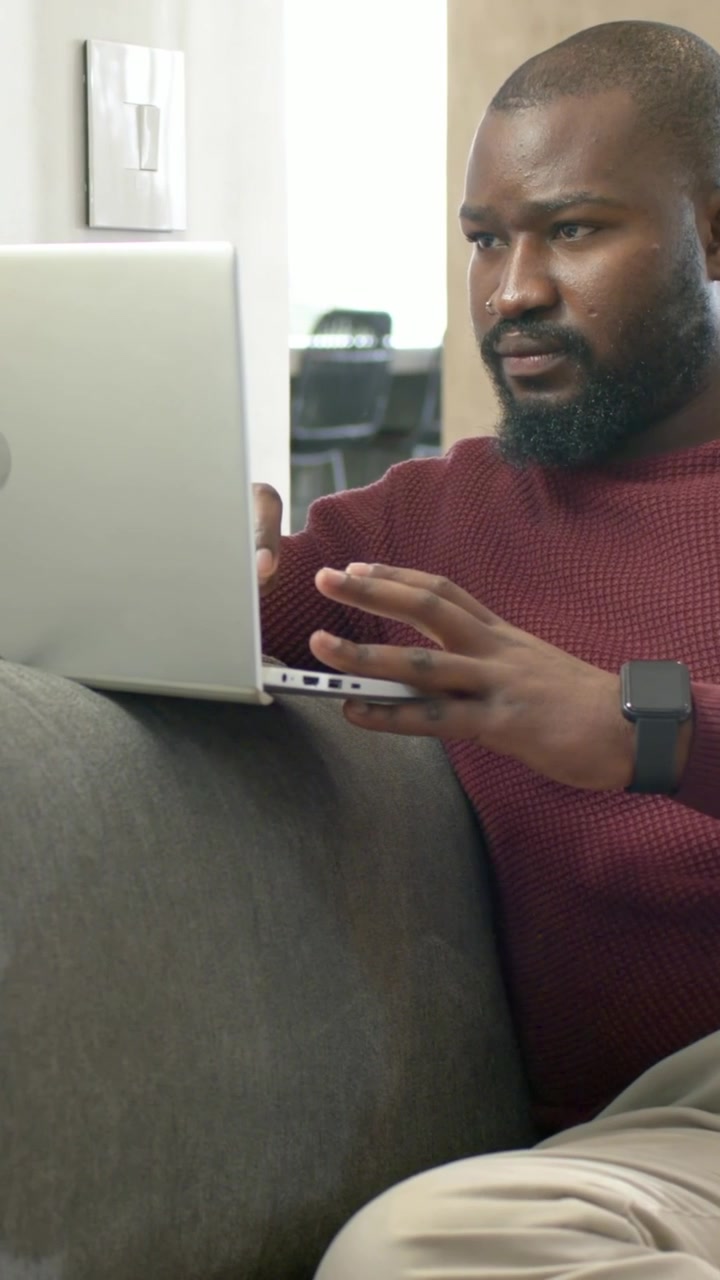 Vertical video: Placing silver laptop, mid-adult man in maroon sweater typing on gray sofa for work