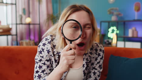 woman holding magnifying glass near face looking into camera with big zoomed funny eye analyzing