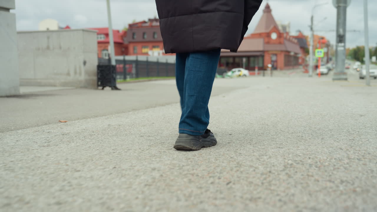 close de pés caminhando em uma praça, clima urbano de trajeto matinal, passos rápidos, detalhes do pavimento, edifícios coloridos ao fundo, bainha do casaco balançando, jeans, sapatos pretos, movimento rítmico, <1>clipe</1> de <2>stock</2> loopável