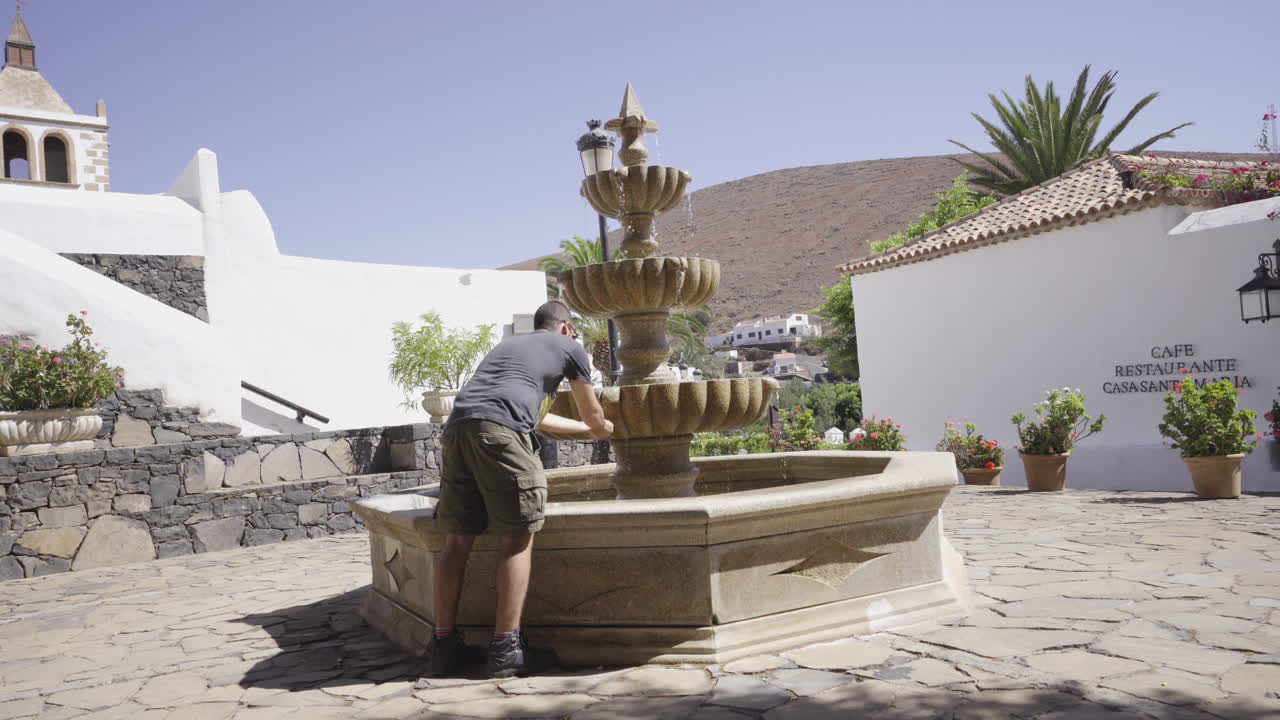 Młody podróżnik z plecakiem na wyspie Fuerteventura (Wyspy Kanaryjskie, Hiszpania) pijący z fontanny w centrum tradycyjnego kamiennego miasteczka w pustynnych górach.