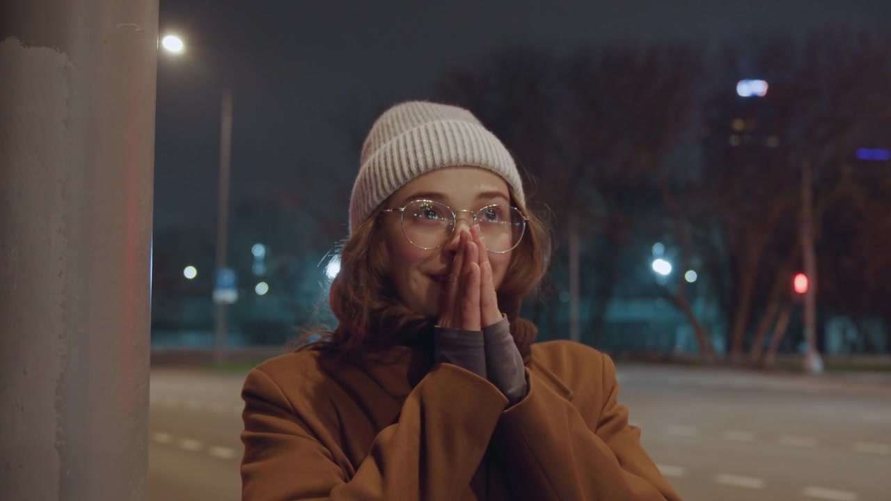 Night street bus stop white woman student waiting then surprised and relieved, city lights and empty roadway, brown coat, beanie and glasses, scarf visible, soft lamppost glow, cold breath in air