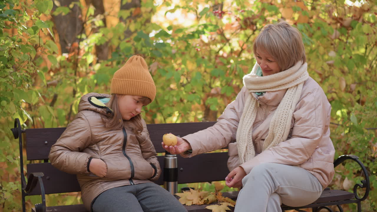 Mother warmly observes daughter seated calmly on park bench in soft autumn sunlight, handing fruit to her with gentle smile, bright leaves surrounding them in tranquil, heartfelt outdoor moment