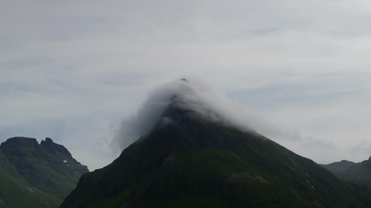 Nuvens baixas cobrem um pico de montanha íngreme em forma de pirâmide em Lofoten, Noruega, sob a suave luz cinzenta do dia.