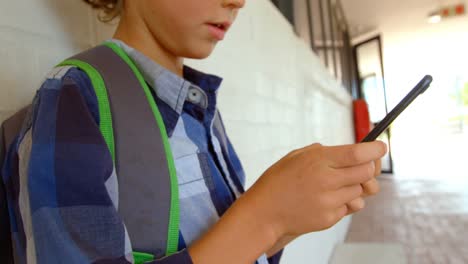 Vista-Lateral-De-Un-Colegial-Caucásico-Usando-Un-Teléfono-Móvil-Mientras-Se-Apoya-En-La-Pared-En-El-Pasillo-De-La-Escuela-4k