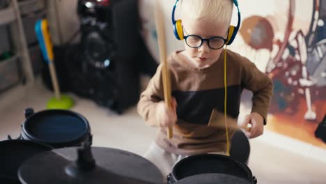 Close-up-shot-from-above-of-a-small-albino-boy-with-white-hair-wearing-blue-glasses-playing-an-electronic-drum-kit-using-special-wooden-sticks-at-home-in-his-room