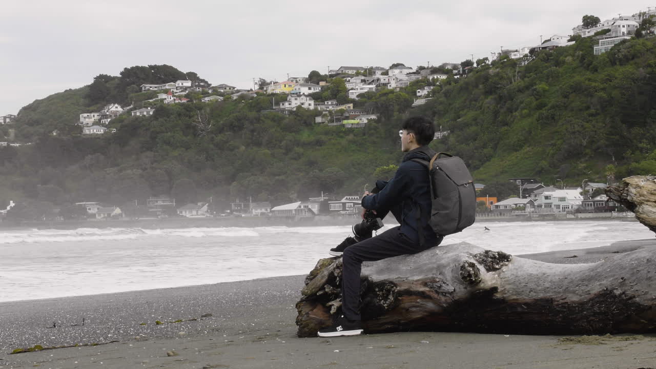 A Malaysian Photographer is Resting on a Fallen Log Along the Seashore at Lyall Bay in Wellington, New Zealand - Medium Shot