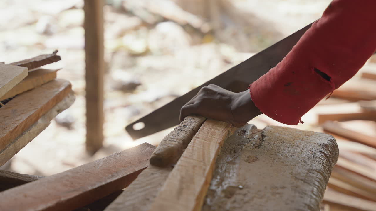 Worker sawing stacked wooden boards on bench, steady hands guiding blade to shape edges and remove excess, maker trimming boards for assembly, warm workshop light and textured wood pile