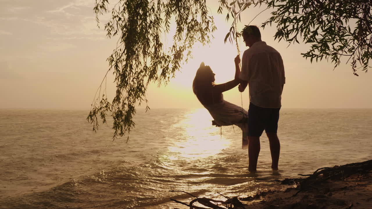 한 남자가 바다를 배경으로 그네를 타고 아내를 굴리고 있는 젊은 부부의 실루엣