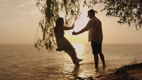 Silhouetten-Eines-Jungen-Paares,-Ein-Mann-Rollt-Seine-Frau-Auf-Einer-Schaukel-Vor-Dem-Hintergrund-Des-Meeres