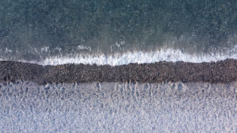 top down aerial view above layered white sand and pebble beach washed over by foaming ocean waves