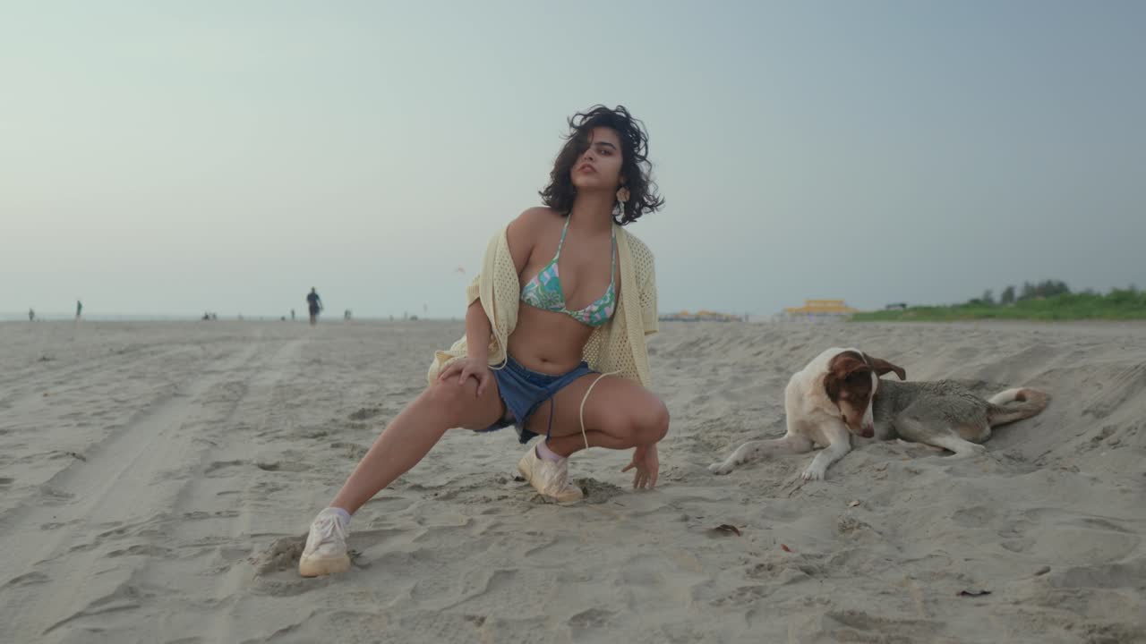 Giovane donna in posa giocosa su una spiaggia sabbiosa con un cane sdraiato accanto a lei, in abbigliamento da spiaggia casual, in una giornata nuvolosa