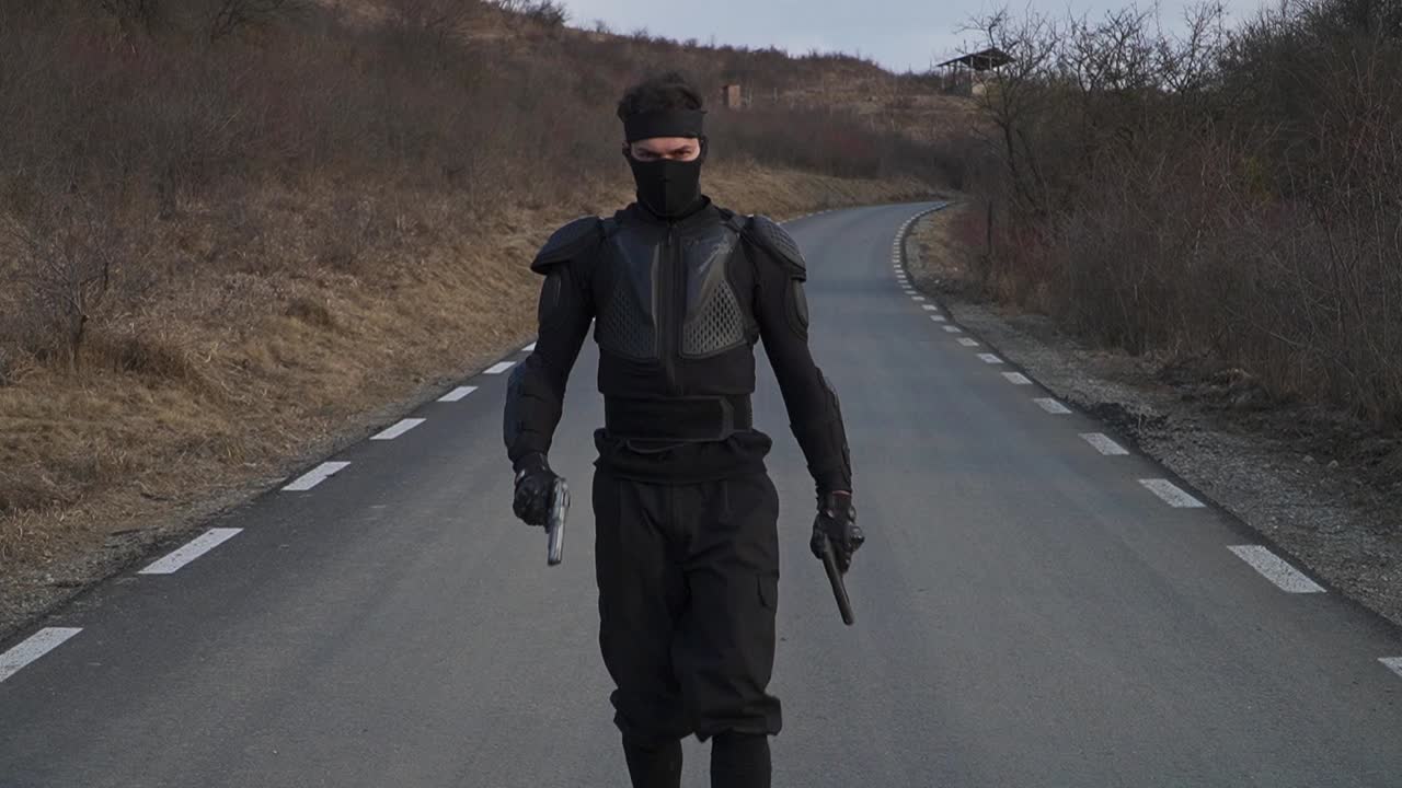 A man dressed in black tactical gear, including a mask and body armor, walks alone on an empty, rural road holding handguns in both hands. The surrounding area is dry with leafless bushes and hills.