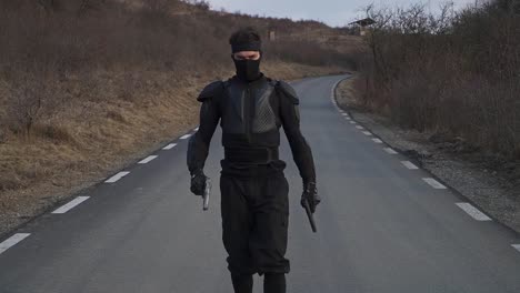 a man dressed in black tactical gear, including a mask and body armor, walks alone on an empty, rural road holding handguns in both hands. the surrounding area is dry with leafless bushes and hills.