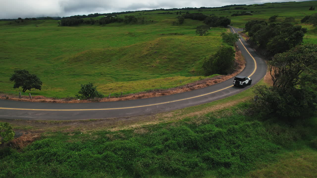 무성한 푸른 언덕과 나무가 있는 경치 좋은 도로를 달리는 흰색 자동차의 드론 샷