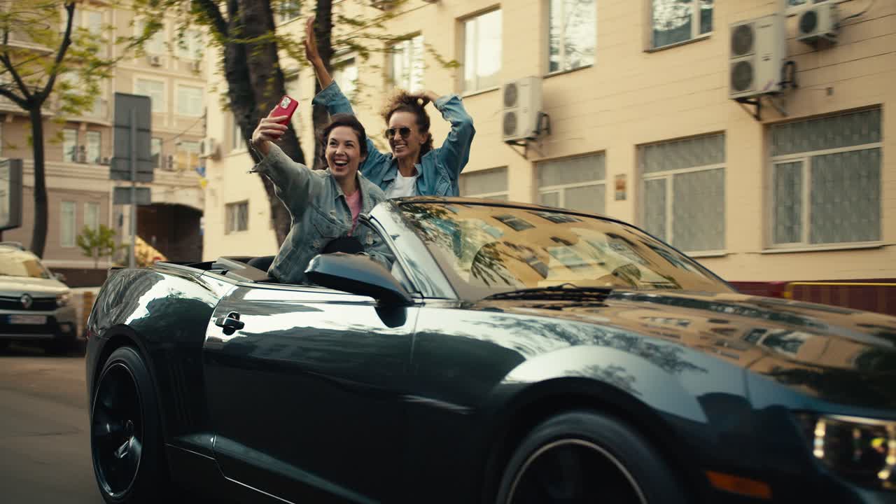 두 명의 행복한 소녀가 회색 컨버터블을 타고 재미있게 셀카를 찍고 있습니다. 도시에서 행복한 청소년
