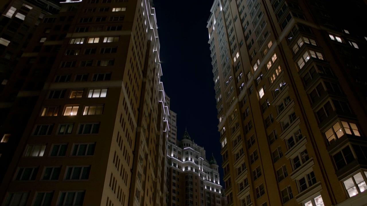 Vista urbana notturna suggestiva tra alti edifici residenziali, con finestre calde che brillano contro un cielo scuro e uno stretto corridoio stradale che crea forte profondità e simmetria.