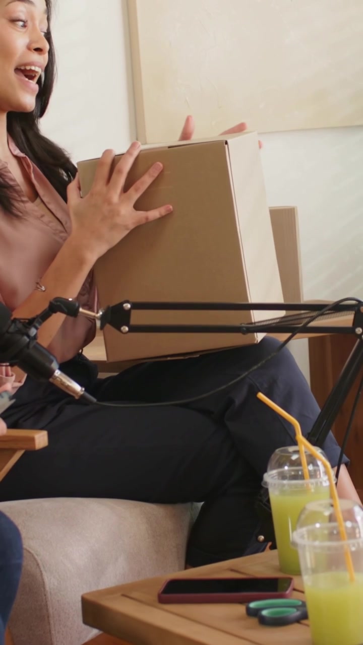 Vertikales Video: Sprechende Frau in pinker Bluse und dunkler Hose greift nach einer Box, um den Inhalt auf dem Sofa in der Nähe eines Mikrofons zu zeigen.