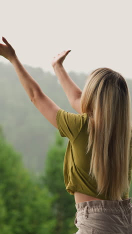 woman with loose hair reaches hands to catch rain drops spending time at highland eco resort on gloomy day backside view slow motion