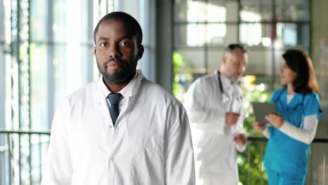 retrato de un médico afroamericano vestido de blanco mirando a la cámara y de pie en la clínica