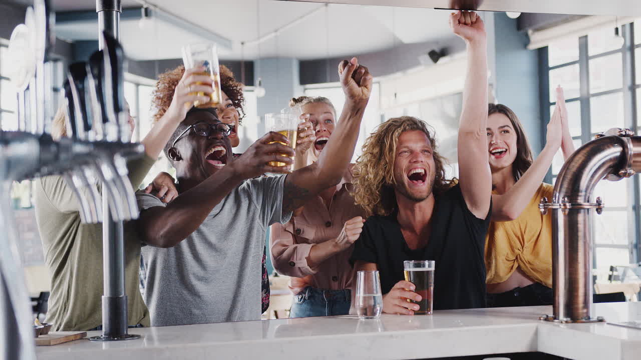 Group Of Male And Female Friends Celebrating Whilst Watching Game On Screen In Sports Bar