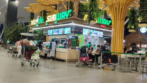bangalore airport passed next to the foodcourt with people standing waiting in line for the subway sandwich store and passengers.