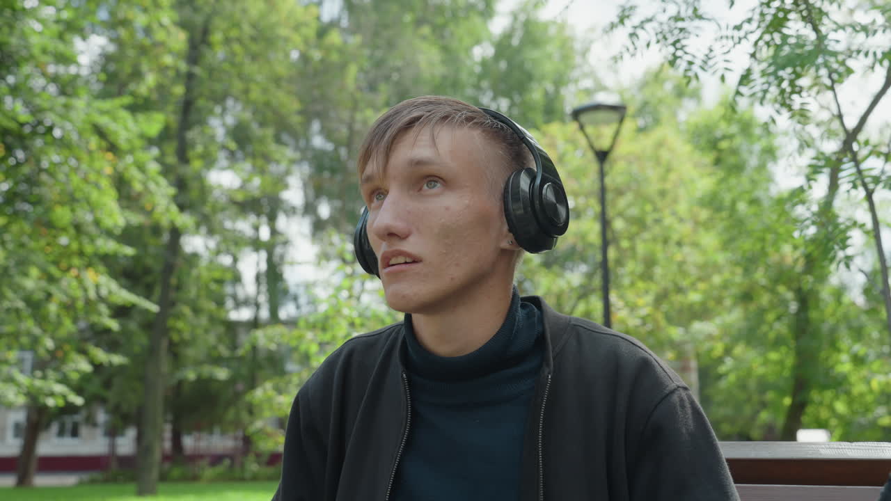 Uomo bianco seduto su una panchina del parco ascolta la musica con cuffie nere, capelli bagnati per la pioggia leggera, sistema i padiglioni e guarda verso gli alberi e il lampione, maglione casual, primo piano spontaneo, umore riflessivo