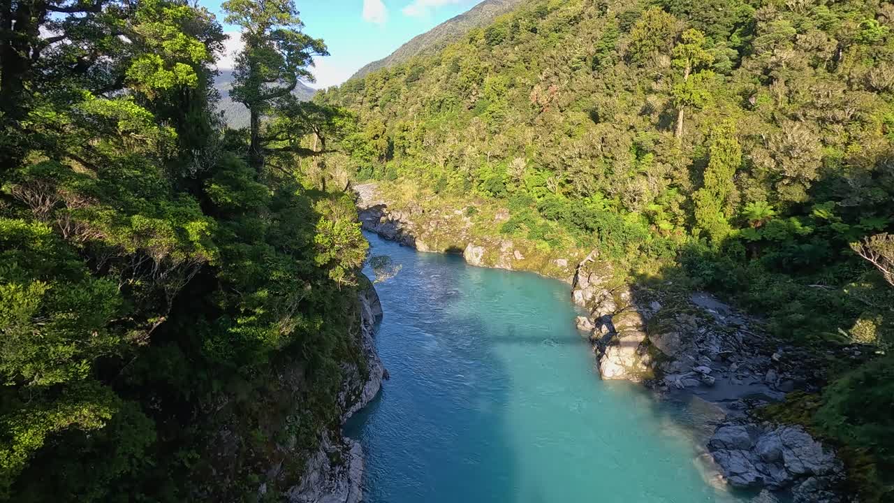 view from a high suspension bridge looking out at the bright blue waters at Hokatika Gorge in New Zealand's South Island. This beautiful blue water is surrounded by cliffs and greenery on either side