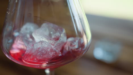 closeup of transparent wine glass filled with red drink and melting ice cubes placed on wooden table as another ice cube is dropped inside under warm daylight creating refreshing visual detail