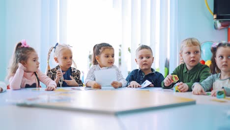 little children studying at the lesson. elementary school children sitting together at the round table and working with paper pictures. small girl gives one card to her teacher.