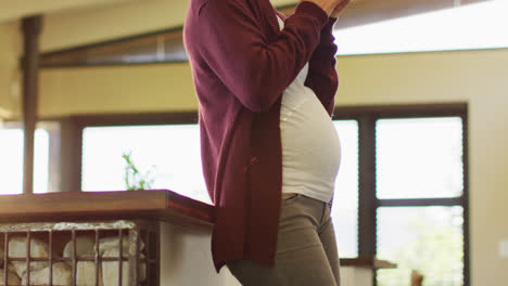 midsection of caucasian pregnant woman standing in kitchen, touching belly and drinking tea