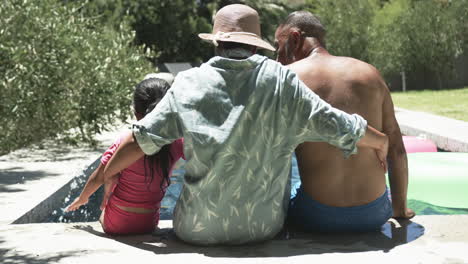 unos abuelos biraciales se sientan con una joven biracial junto a una piscina, disfrutando del sol.