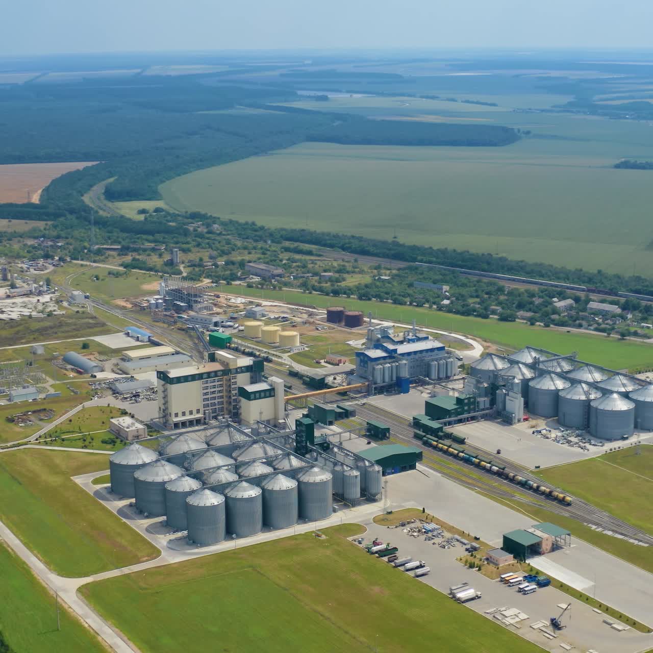 Large industrial territory among beautiful nature. Modern industry for keeping and processing crop. Agricultural complex with grain elevators on field in summer. Aerial view