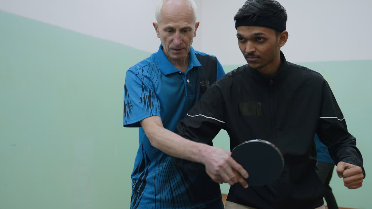 Coach carefully demonstrates proper standing position to student during tennis training, guiding arm and paddle placement with focused attention in indoor practice setting