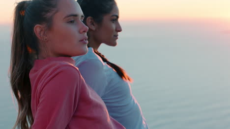 Amigas-Sentadas-En-La-Cima-De-La-Montaña-Mirando-La-Tranquila-Vista-Del-Océano-Al-Atardecer-Dos-Mujeres-Descansando-Después-De-Una-Caminata-Disfrutando-De-Una-Tranquila-Aventura-De-Viaje-Al-Aire-Libre