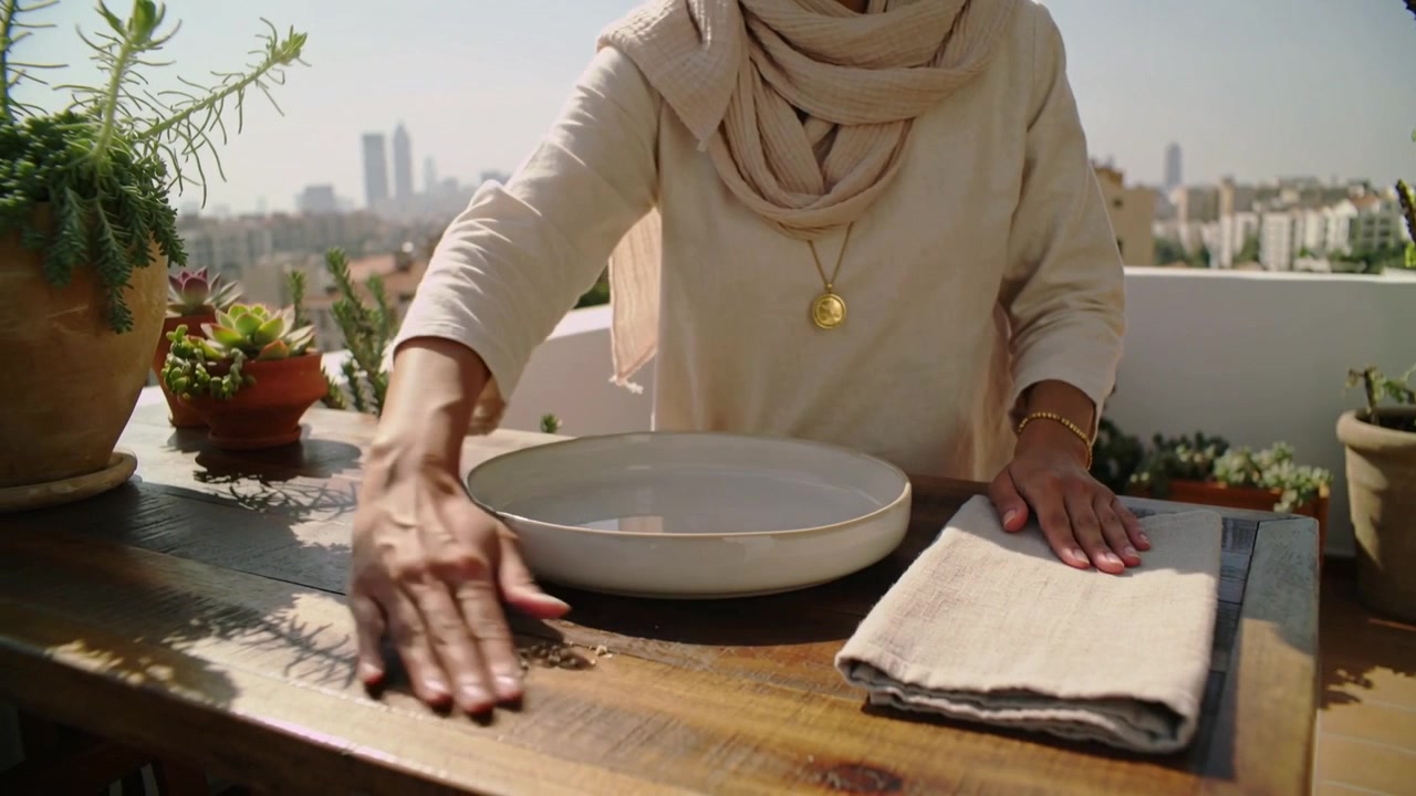 Donna con sciarpa beige che prepara, sistema uno strofinaccio di lino, lo piega e pulisce le briciole sul balcone.