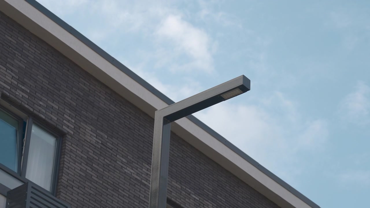 Low-angle exterior view of a modern brick building with windows, metal roofline detail, and a streetlight set against a partly cloudy sky. The composition