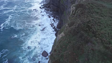 Drohne-Erfasst-Den-Rand-Des-Hügels-In-Isla-Und-Auch-Das-Blaue-Kantabrische-Meer,-Das-Aus-Großer-Höhe-Auf-Die-Felsen-Trifft