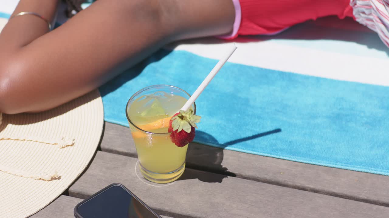 Afroamerikansk kvinne soler seg under sollyset, i rød badedrakt, hviler på et håndkle på terrassen med en drink.