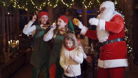 fairy characters having fun at christmas. little girl singing and dancing together with santa claus and happy elves in the decorated room.