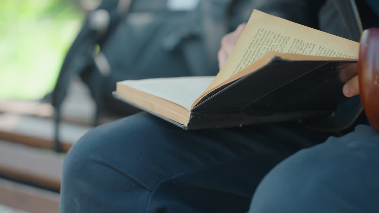 Homme sur un banc de parc lisant un carnet, les mains feuilletant les pages en gros plan. Un étudiant décontracté est assis avec un sac à dos sur des lattes de bois, un pantalon en denim, la lumière du soleil filtrant, une ambiance calme et réfléchie, du papier texturé, page lente