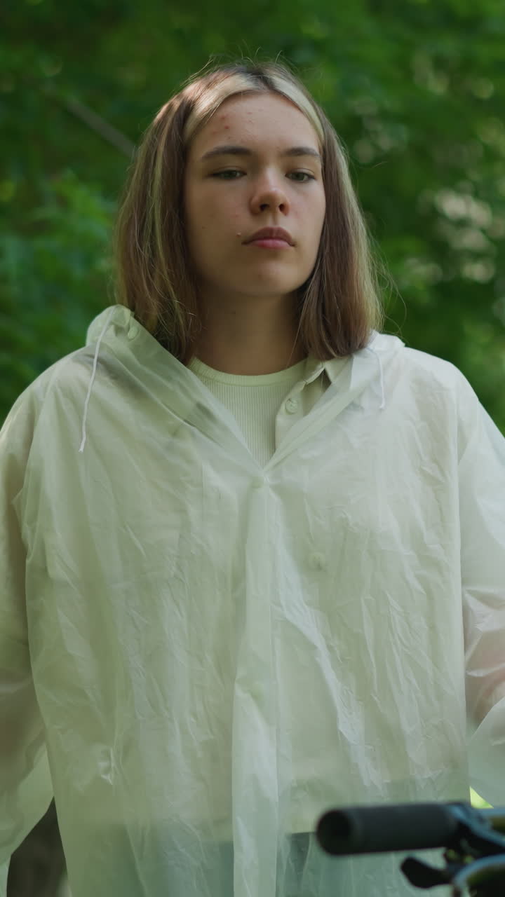 반투명한 레인코트를 입은 젊은 여성은 생각에 빠져 서 있고, 손은 자전거 손잡이에 혀 있으며, 초록색과 나무로 둘러싸여 있으며, 그녀는 멀리서 상적으로 쳐다보고 있습니다.