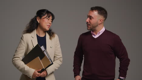 Studio-Portrait-Of-Smiling-Male-And-Female-Teachers-Walking-Towards-Camera-And-Talking-Against-Grey-Background