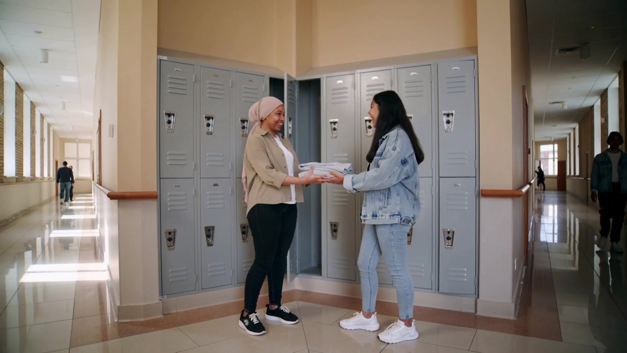 Stepping right student in denim handing stack to left friend in hijab, lockers lining school hall
