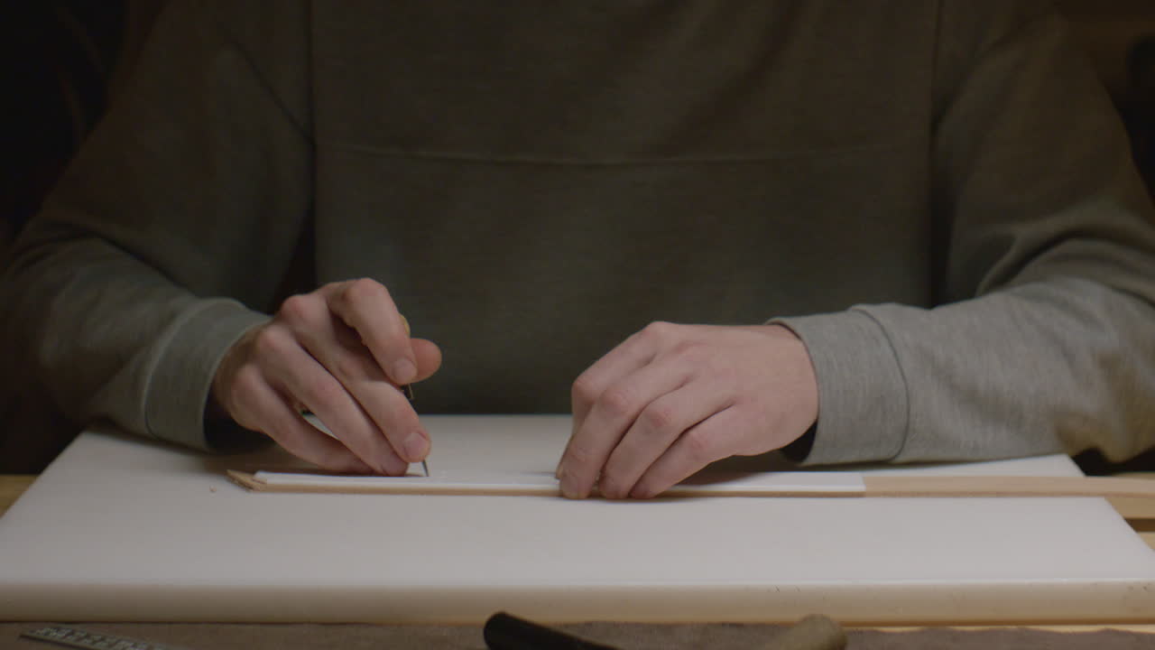A craftsman uses an awl to carefully mark a leather strip for precise cutting in a workshop setting.