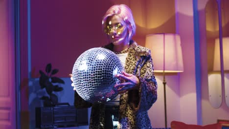 young stylish woman in glasses holding disco ball in her hands and looking at camera in a retro party at home